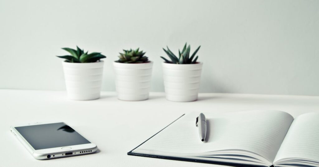 Three White Ceramic Pots With Green Leaf Plants Near Open Notebook With Click Pen on Top